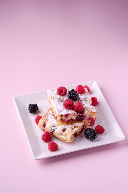 Homemade piece of cake, two pie slices with raspberry and blackberry berries, powdered sugar, in white square plate on pink 