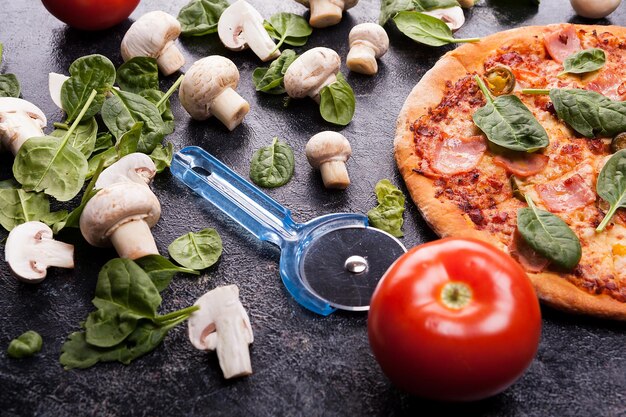 Homemade pizza next to mushrooms, baby spinach and tomatoes with a pizza cutter on dark wooden background