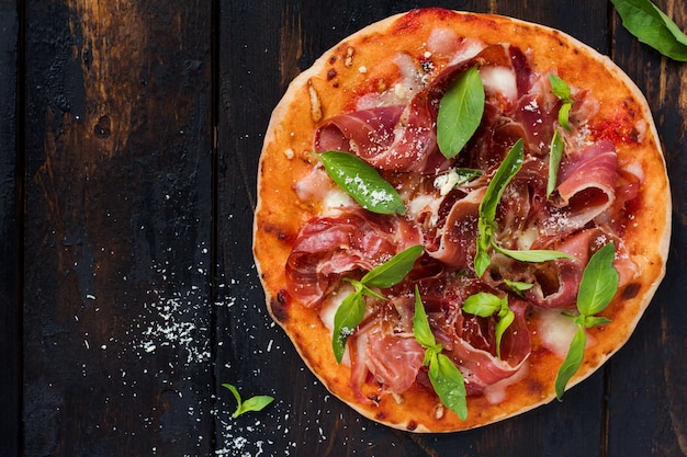 Homemade pizza with jamon, mozzarella and basil leaves on an old wooden background. Top view.