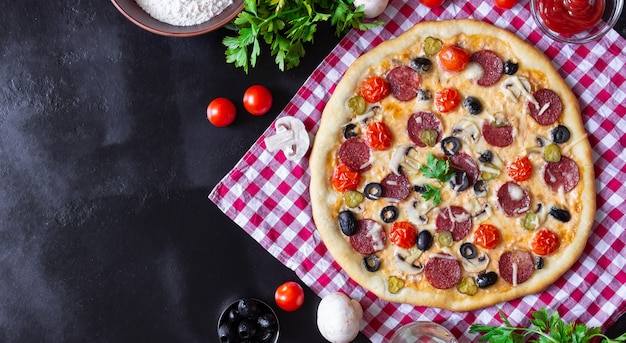 Homemade pizza with salami, mushrooms and cherry tomatoes on a black background. A red checkered towel. Free space for text, top view.