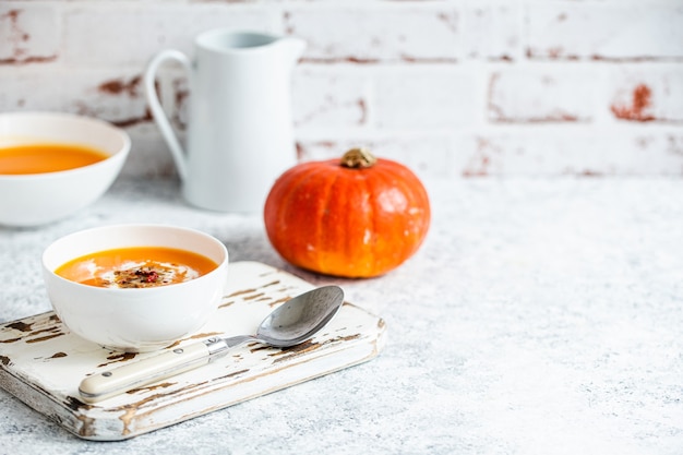 사진 홈메이드 호박 크림 수프는 전체 호박, 각도 전망, 선택적 초점으로 장식된 스푼이 있는 흰색 테이블에 있는 흰색 세라믹 그릇에 제공됩니다. 가을의 안락한 음식, 텍스트를 위한 배경 공간