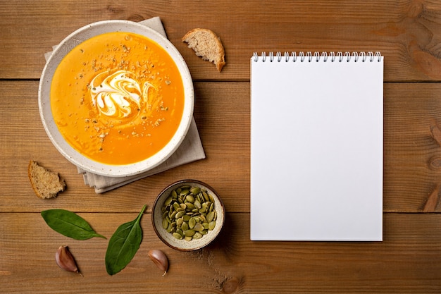 Homemade pumpkin soup in white bowl with notepad flat lay on brown wooden background with copy space.
