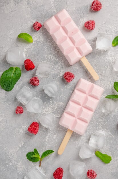 Gelato al lampone fatto in casa o ghiaccioli decorati con foglie di menta verde e frutti di bosco congelati con ghiaccio su uno sfondo grigio cemento.