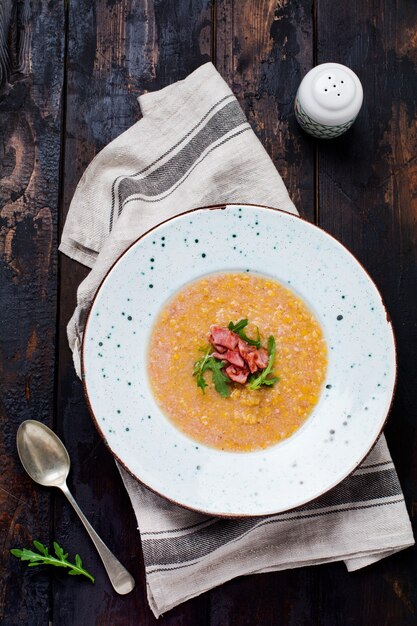 Homemade red lentil cream soup with bacon and arugula in a white plate on an old wooden surface. Rustic style. Top view.