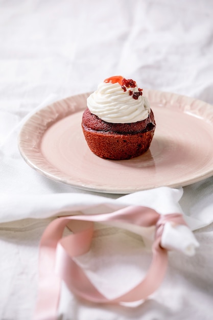 Домашний красный бархатный кекс со взбитыми сливками на розовой керамической тарелке, белая салфетка с лентой на белой льняной скатерти