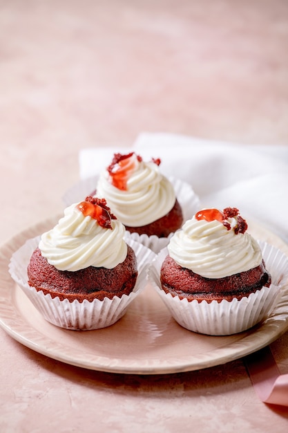 Cupcakes di velluto rosso fatti in casa con panna montata sul piatto in ceramica rosa, tovagliolo bianco con nastro sul tavolo di consistenza rosa.