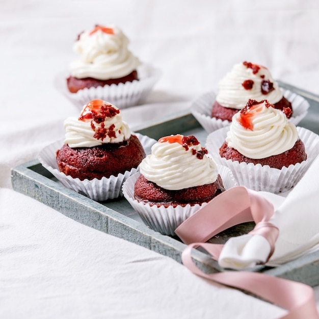 Homemade Red velvet cupcakes with whipped cream on wooden tray, white napkin with ribbon on white linen table cloth. Square image