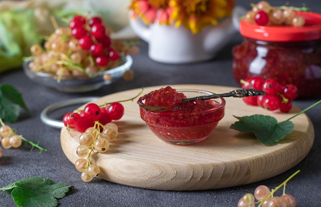 Homemade red and white currant jam in transparent bowl on a wooden board as well as jar of jam in background