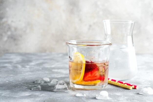 Limonata fredda rinfrescante fatta in casa da bacche e frutti di limone in un bicchiere con cubetti di ghiaccio su un tavolo di marmo grigio.