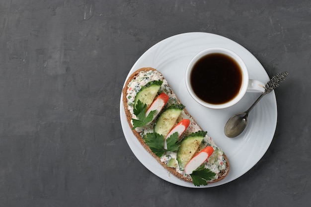 Homemade sandwich with crab sticks, cucumber, cream cheese and a cup of coffee on dark surface