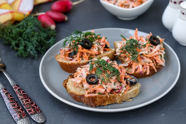 Homemade sandwiches with carrots and radishes, decorated with boiled egg and black olives in a plate against a dark 