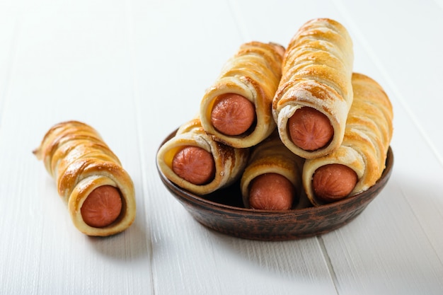 Homemade sausage rolls neatly stacked on a clay bowl on a white table.