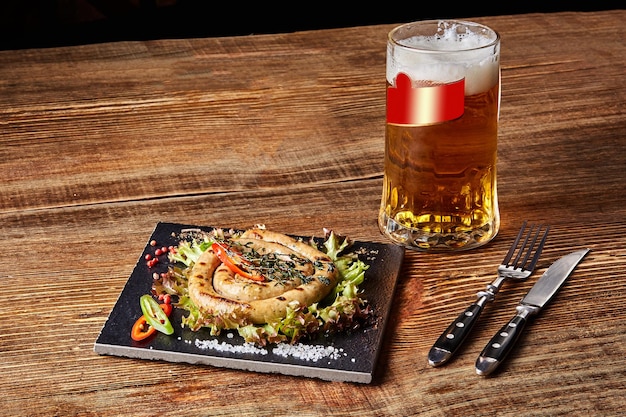 슬레이트 접시에 마늘과 향신료를 곁들인 홈메이드 소시지 나무 테이블에 있는 접시 뒤에 있는 맥주 한 잔