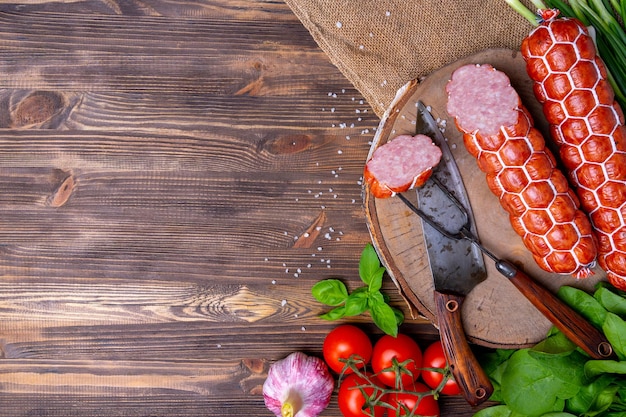 Домашняя колбаса с овощами на деревянном фоне, вид сверху. Нож и вилка с кусочком колбасы