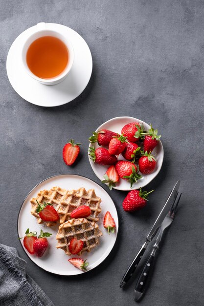 Homemade soft waffles with fresh strawberries and powdered sugar in a plate on a dark background with a cup of tea Traditional Belgian waffles Healthy breakfast