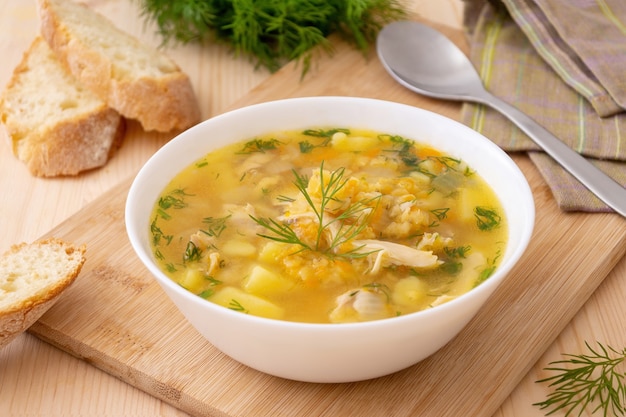 Homemade soup with red lentils and chicken, garnished with dill in a white bowl, served with fresh bread on a wooden cutting board.