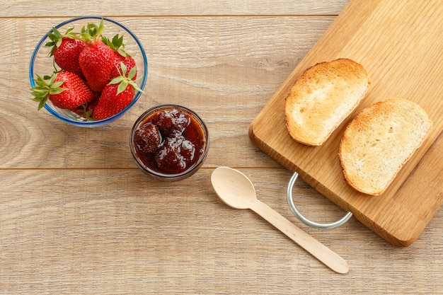 Homemade strawberry jam and fresh berries in glass bowls, toasts on cutting board with spoon on wooden desk. Top view.