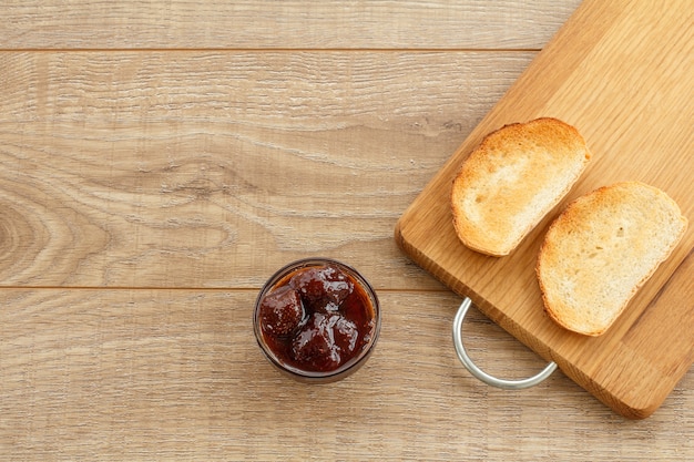 Homemade strawberry jam in a glass bowl and toasts on cutting board on wooden desk. Top view.