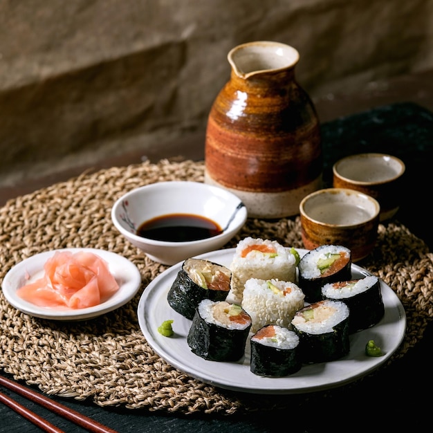 Homemade sushi rolls set with salmon japanese omelette avacado and soy sauce with chopsticks on straw napkin over balck wooden table Ceramic drink sake set Japanese style dinner Square image