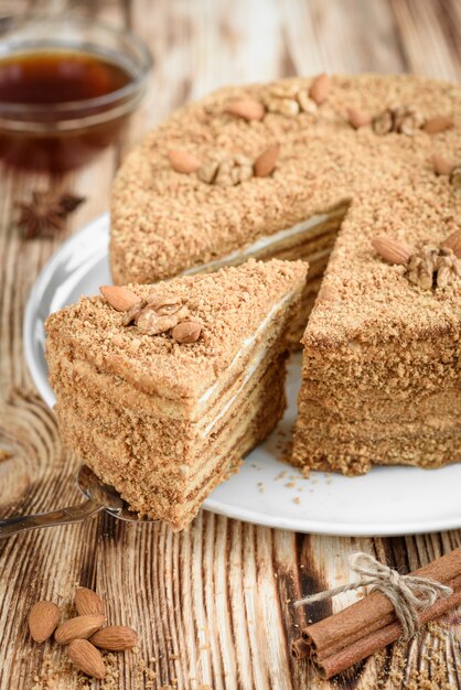 Homemade sweet layered honey cake with nuts on white plate on wooden table with bowl of honey, cinammon and badian.