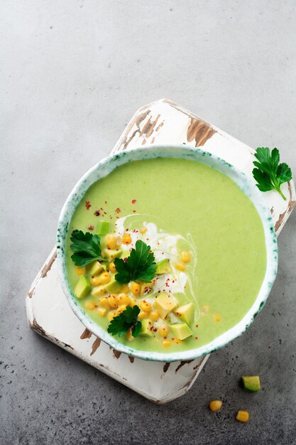 Homemade tender soup puree from avocado and corn with cream in rustic ceramic plate on gray concrete old surface. Selective focus. Top view.