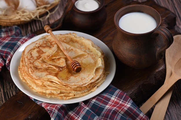 Homemade thin pancakes with honey stacked in a stack, on a wooden table with a mug of milk, a pot of sour cream and eggs in a basket. Traditional Slavonian, pagan holiday (Maslinitsa). Country style f