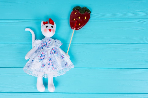 Giocattolo fatto in casa a forma di gatto con un vestito che tiene in mano una lecca-lecca colorata come una fragola su una tavola di legno turchese