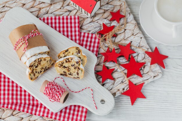 Homemade traditional christmas dessert stollen with dried berries, nuts and powdered sugar on top stands on white rustic wooden table with cup of coffee.