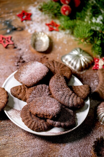 Домашнее традиционное рождественское песочное печенье шоколадные полумесяцы с сахарной пудрой какао в керамической тарелке с формами для печенья, елкой, красными рождественскими звездами, украшениями на деревянном фоне