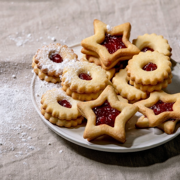 Homemade traditional Linz shortbread biscuits cookies with red jam and icing sugar on ceramic plate over linen tablecloth. Square image