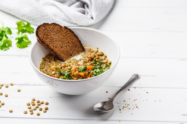 Homemade vegan lentil soup with vegetables, bread and cilantro, white wooden background. 