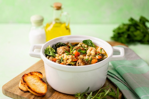 Homemade vegetable soup with meatballs and spinach in a white saucepan on a green background