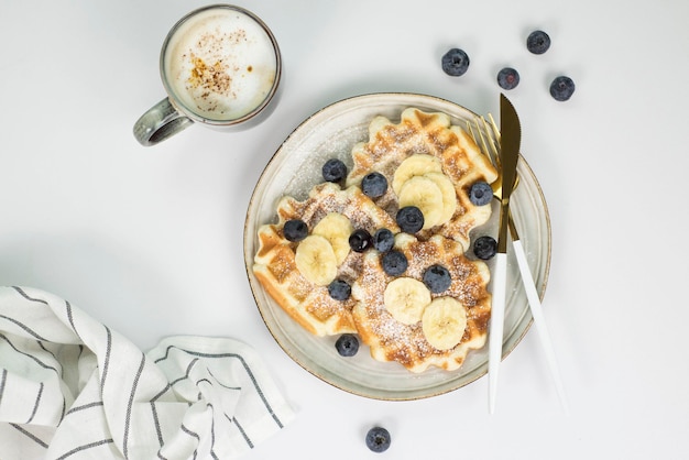 Homemade viennese waffles with blueberries and banana and cup of coffee on a light background