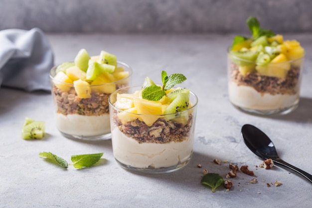 Foto parfait di yogurt fatto in casa con muesli, kiwi, ananas e noci in un bicchiere per una sana colazione su sfondo concreto