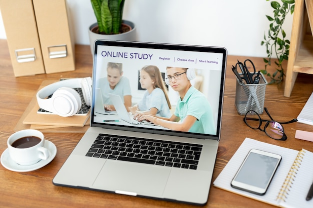 Homepage of educational website on laptop display surrounded by cup of coffee, smartphone and other supplies