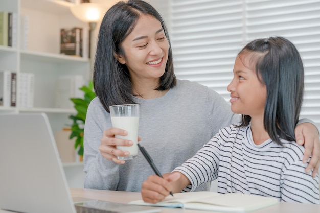 Homeschool Asian young little girl learn read and do homework with kind mother help teach and encourage Mom pass on a glass of milk to daughter Girl happy to study and education together with mom