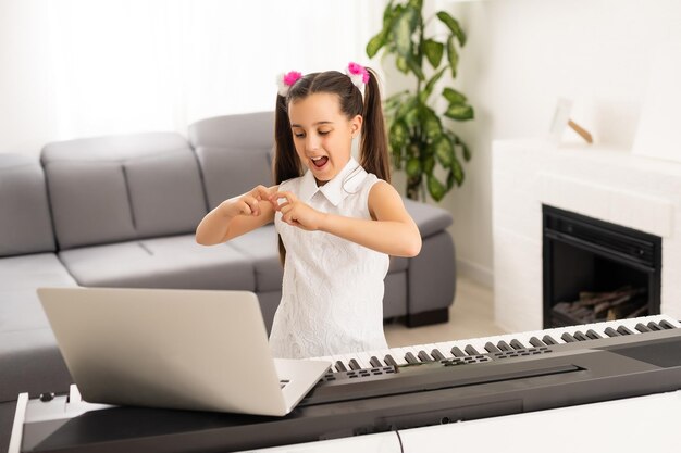 Маленькая девочка, обучающаяся на дому, учится играть на фортепиано с компьютера, подключаясь к онлайн-классу интернет-музыки школьным учителем. Новый нормальный образ жизни и образование, концепция обучения студентов дома.