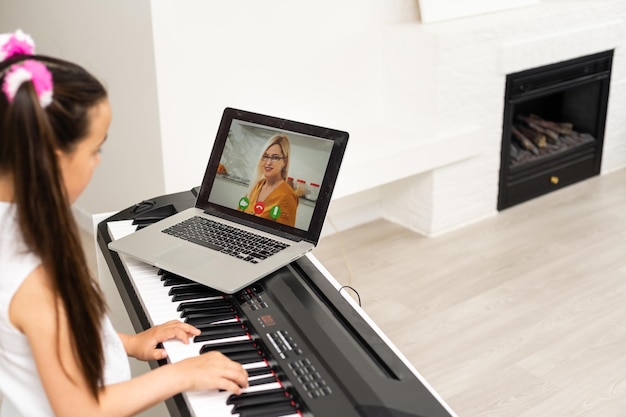 写真 学校の先生によるインターネット音楽オンラインクラスに接続するコンピュータからピアノを学ぶホームスクーリングの小さな幼い子供の女の子。新しい通常のライフスタイルと教育、自宅での学生の勉強のコンセプト。