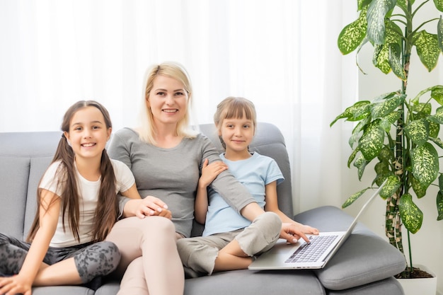インターネットをオンラインで学んでいるホームスクーリングの若い2人の娘は、母親の助けを借りてコンピューターを使用して宿題をし、教え、励まします。家で一緒に教育を勉強して幸せな女の子と妹。