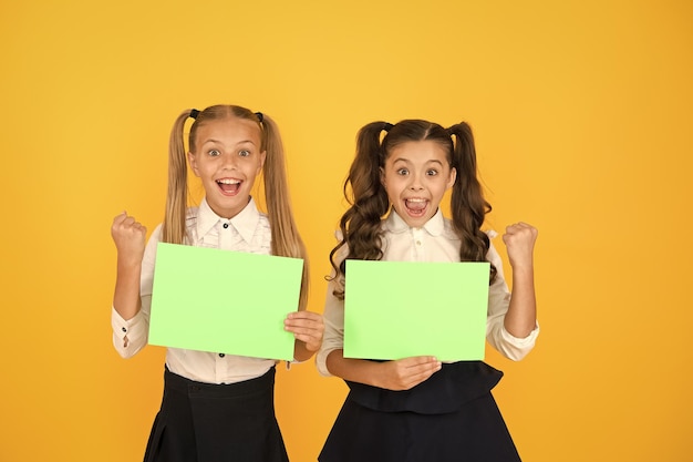 写真 宿題をやった 幼い子供たちが黄色い背景に空の宿題シートを握っている 素敵な女の子たちが空の緑色の学校ポスターで微笑んでいる 宿題のコピースペース