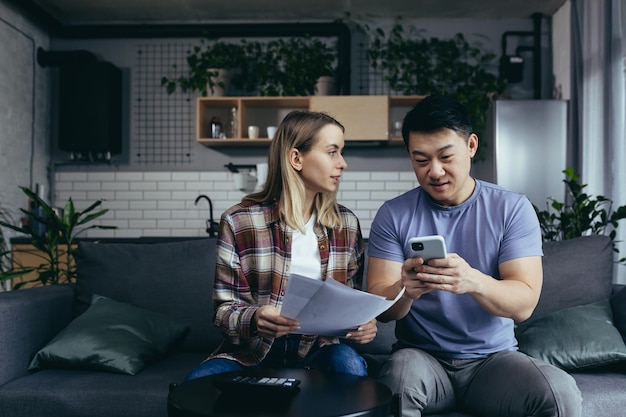 Homework paper multiracial couple man and woman pay bills by phone and app sit at home on the couch happy and smiling