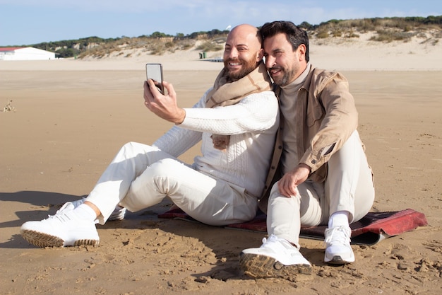 Homoseksueel paar die selfie op strand nemen. Glimlachende homo's van middelbare leeftijd die op zand zitten en samen poseren. Digitale nomade en relatieconcept