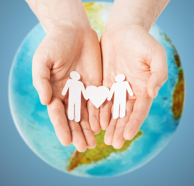 homosexuality, people, love and tolerance concept - close up of male hands holding gay couple paper men with heart shape over earth globe and blue background