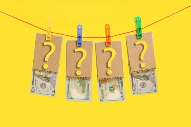 Honderd-dollarbiljetten en vraagtekens worden opgehangen door wasknijpers op een gele achtergrond. Concept van zaken en financiën.