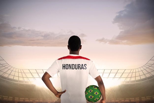 Футболист Гондураса держит мяч против большого футбольного стадиона под облачным голубым небом