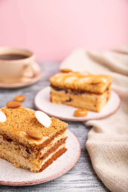 Honey cake with milk cream, caramel, almonds and a cup of coffee on a gray and pink background and linen textile. Side view, close up, selective focus.