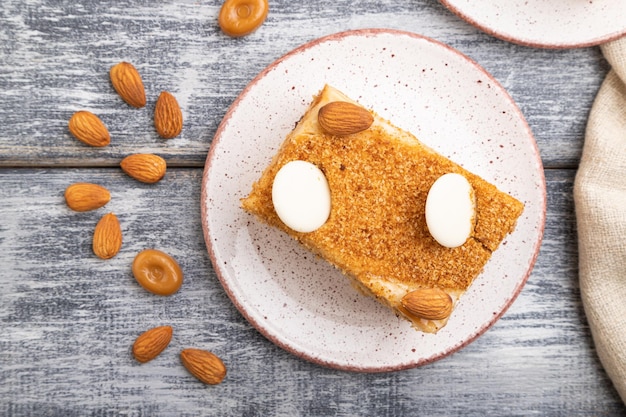 Honey cake with milk cream, caramel, almonds and a cup of coffee on a gray wooden background and linen textile. Top view, flat lay, close up.