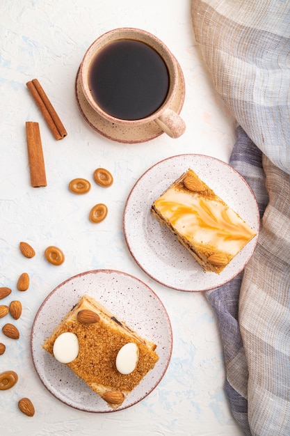 Honey cake with milk cream, caramel, almonds and a cup of coffee on a white concrete background and linen textile. Top view, flat lay, close up.