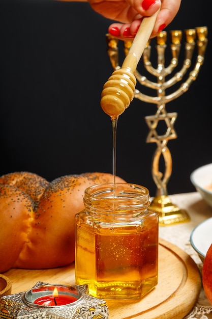 Honey drips from a wooden spoon into a jar next to the menorah challah and pomegranate