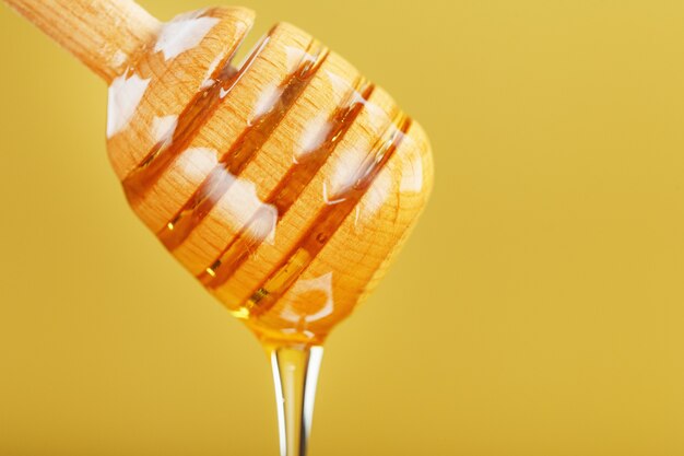 Honey drips in a thin stream from a honey dipper on a yellow background.